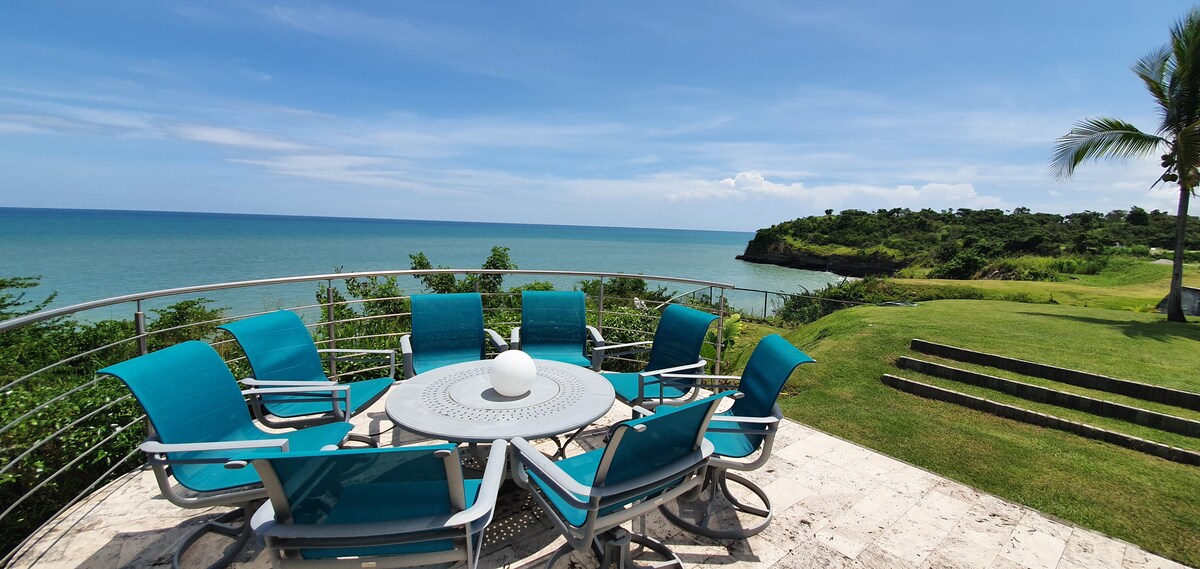 A spacious outdoor terrace features a circular dining table surrounded by eight teal lounge chairs, positioned to overlook the ocean view. Lush greenery and a rocky coastline complement the serene atmosphere, creating a relaxing space for outdoor gatherings.