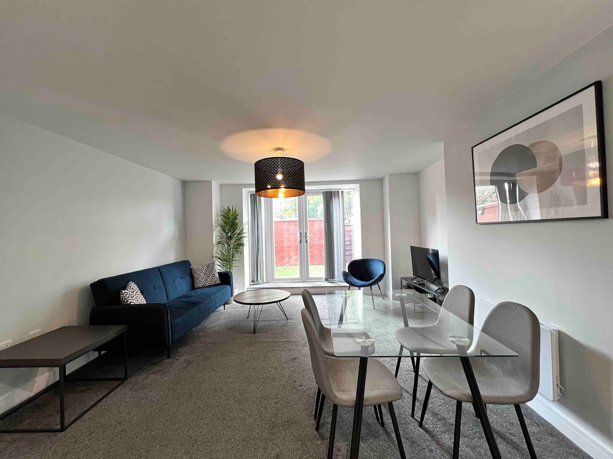 An open plan living and dining area features a comfortable blue sofa, glass dining table with four grey chairs, and a smart TV. Large windows allow natural light to fill the space, and a stylish pendant lamp hangs overhead. A potted plant adds a touch of greenery.