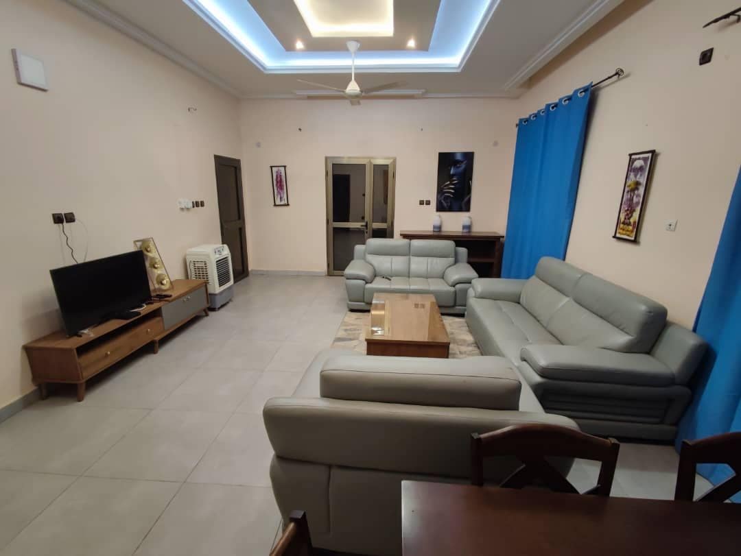 A spacious living room is presented with two large, comfortable sofas arranged around a coffee table. A television sits atop a low wooden cabinet. Bright blue curtains frame the windows, while a fan is mounted on the ceiling, offering a cool atmosphere.