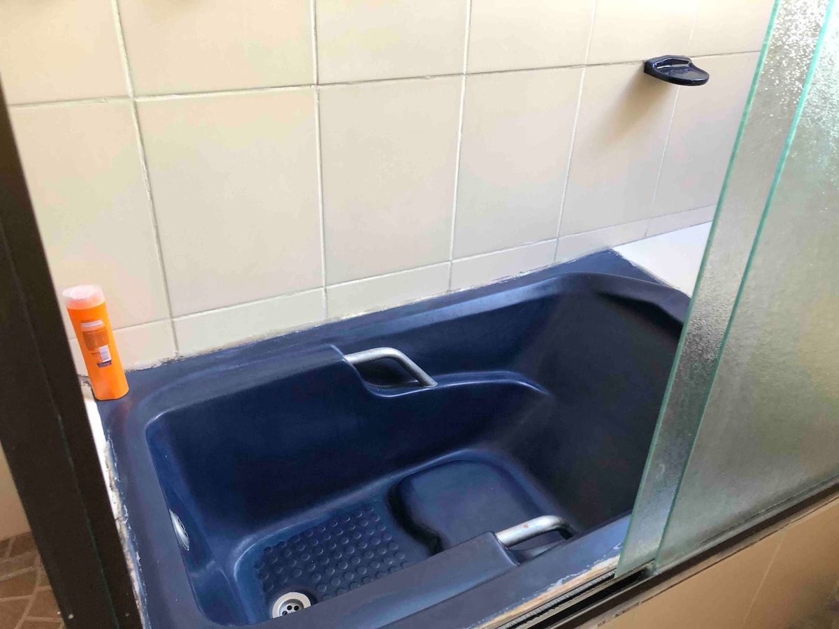 A modern bathtub is set against light tiles, featuring a sleek design with a textured bottom for stability. A showerhead is mounted on the wall nearby, and a bottle of shampoo is positioned on the edge of the tub.
