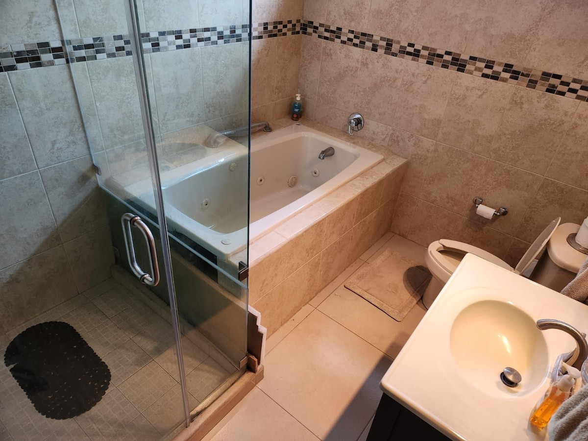 A spacious bathroom features a glass-enclosed shower alongside a modern jetted tub. The elegant tile work includes a decorative border, and natural light enhances the neutral tones. A sink with contemporary fixtures is positioned near a towel, creating an organized and functional space.