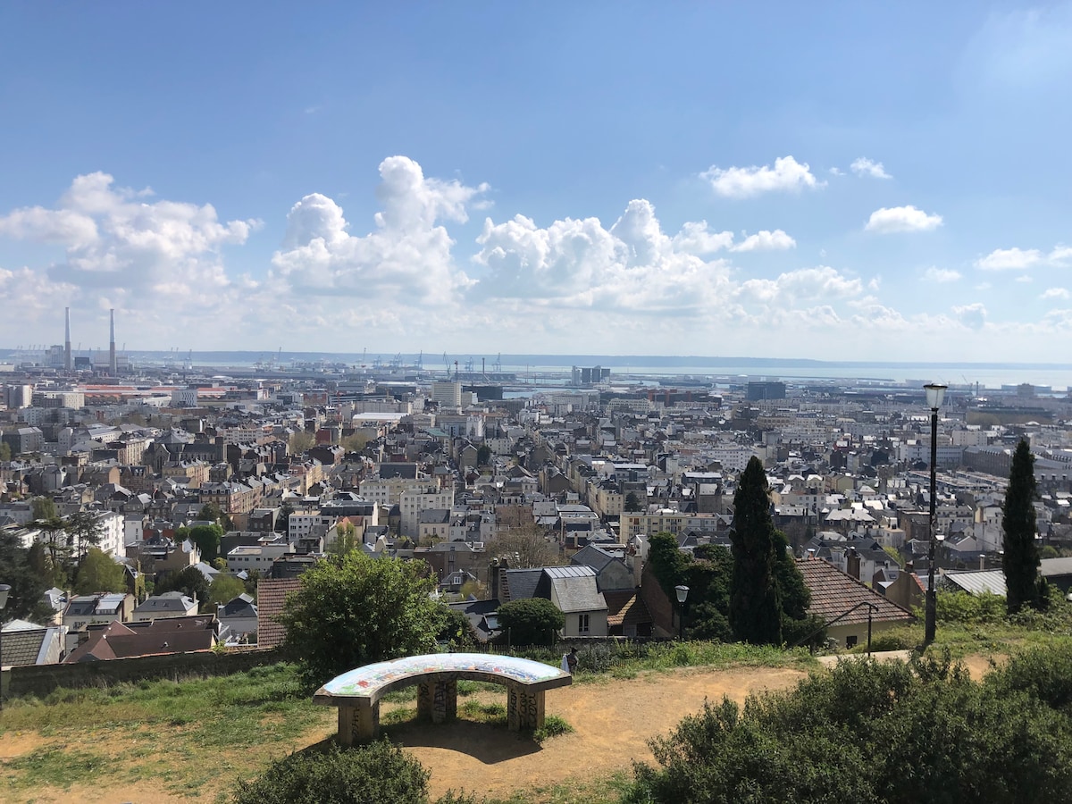A panoramic view of the city and port of Le Havre is presented, featuring a mix of residential buildings and green spaces. In the foreground, a curved bench is visible, inviting relaxation while admiring the expansive skyline and distant harbor under a bright sky.