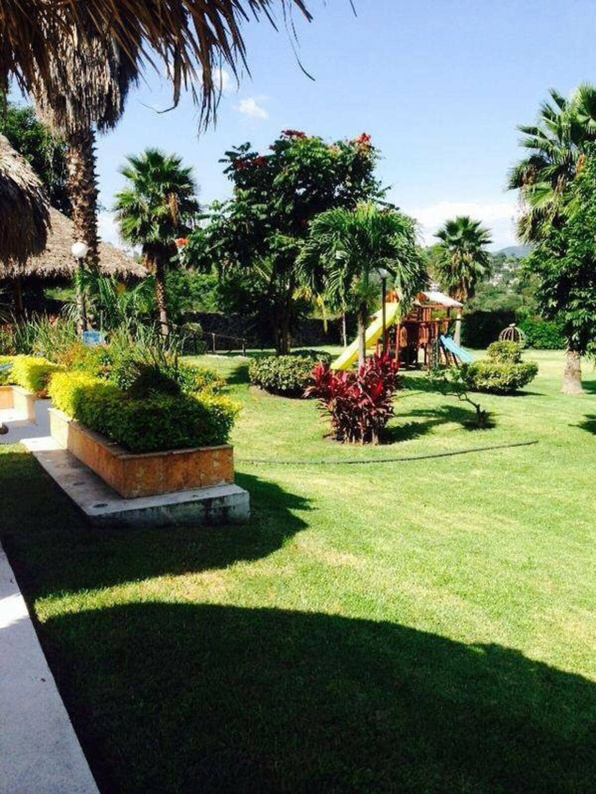 A spacious garden is featured, showcasing lush greenery and vibrant plants. A playground area with slides and swings can be seen in the background. The scene is complemented by palm trees and tropical foliage, enhancing the relaxing outdoor environment.