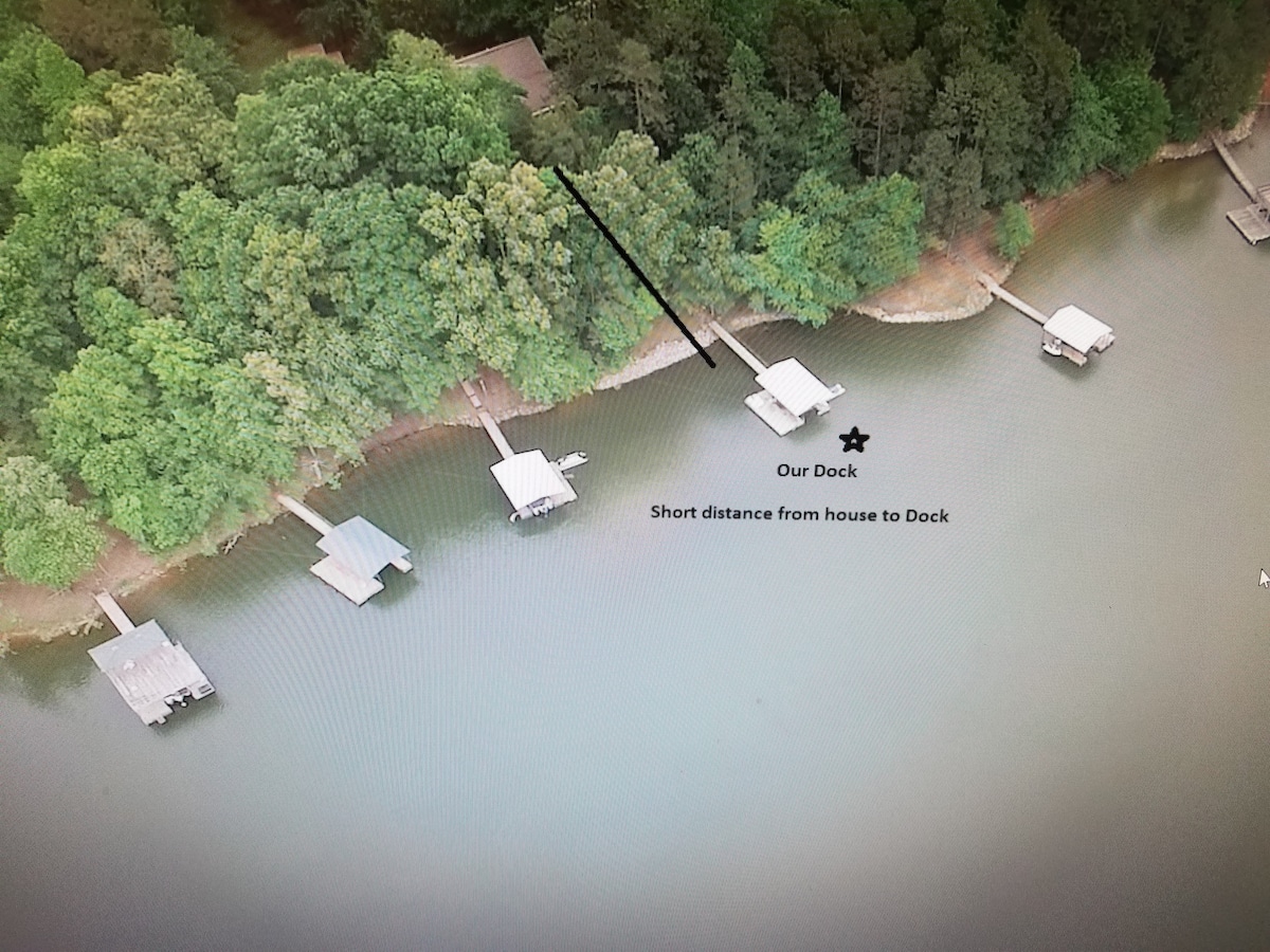 An aerial view of the property reveals a serene lakeside setting with lush green trees bordering the water. A clearly marked dock is highlighted, showcasing a short, accessible path from the house. Several additional docks can be seen along the shore, emphasizing the tranquil environment.