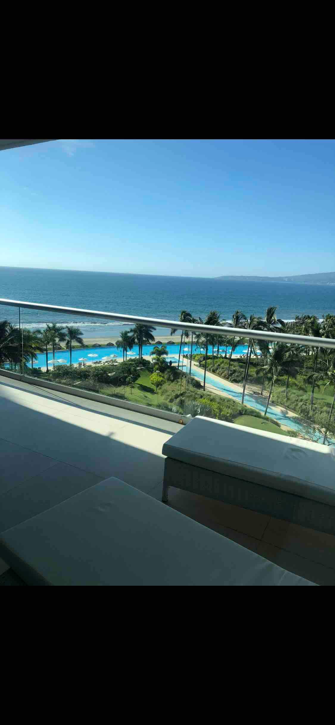 A panoramic view captures the serene ocean waters and lush palm trees from the balcony. A pool area with meandering shapes complements the landscape, visible under clear skies and vibrant sunlight.