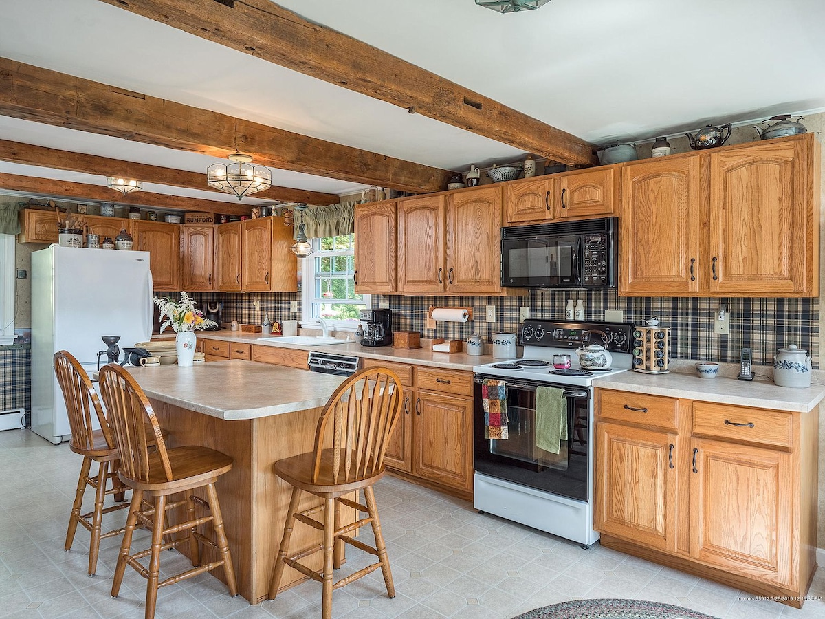 The spacious kitchen features wooden cabinetry and ample counter space. A central food prep island is surrounded by four wooden chairs. Basic cookware and utensils are visible, along with modern appliances, including a refrigerator and microwave, set against a patterned backsplash.