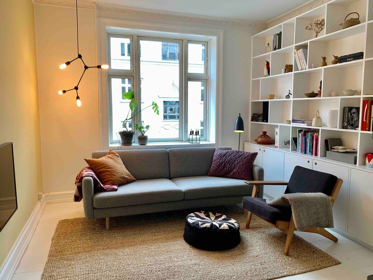 A stylish living area is presented with a gray sofa complemented by colorful pillows. A cozy chair is positioned beside a lamp. A large window allows natural light to illuminate the space, while a bookshelf displays various decorative items and books. A woven rug adds texture to the floor.
