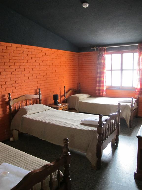 A cozy room features three spacious beds arranged in a triangular layout, with light-colored bedding and wooden frames. An orange brick wall adds warmth to the space, complemented by a window dressed in checkered curtains. A bedside table is positioned next to one of the beds.