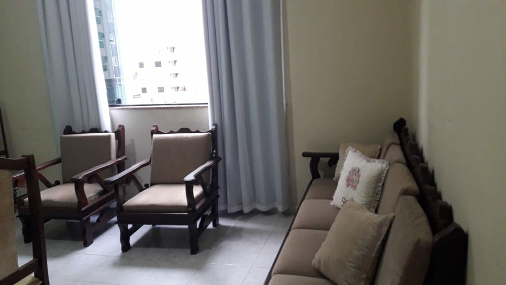 A living area is furnished with a combination of two armchairs and a sofa, all crafted from dark wood. Neutral-toned seat cushions add comfort. Natural light filters through a large window covered by white curtains, illuminating the room's calming atmosphere.