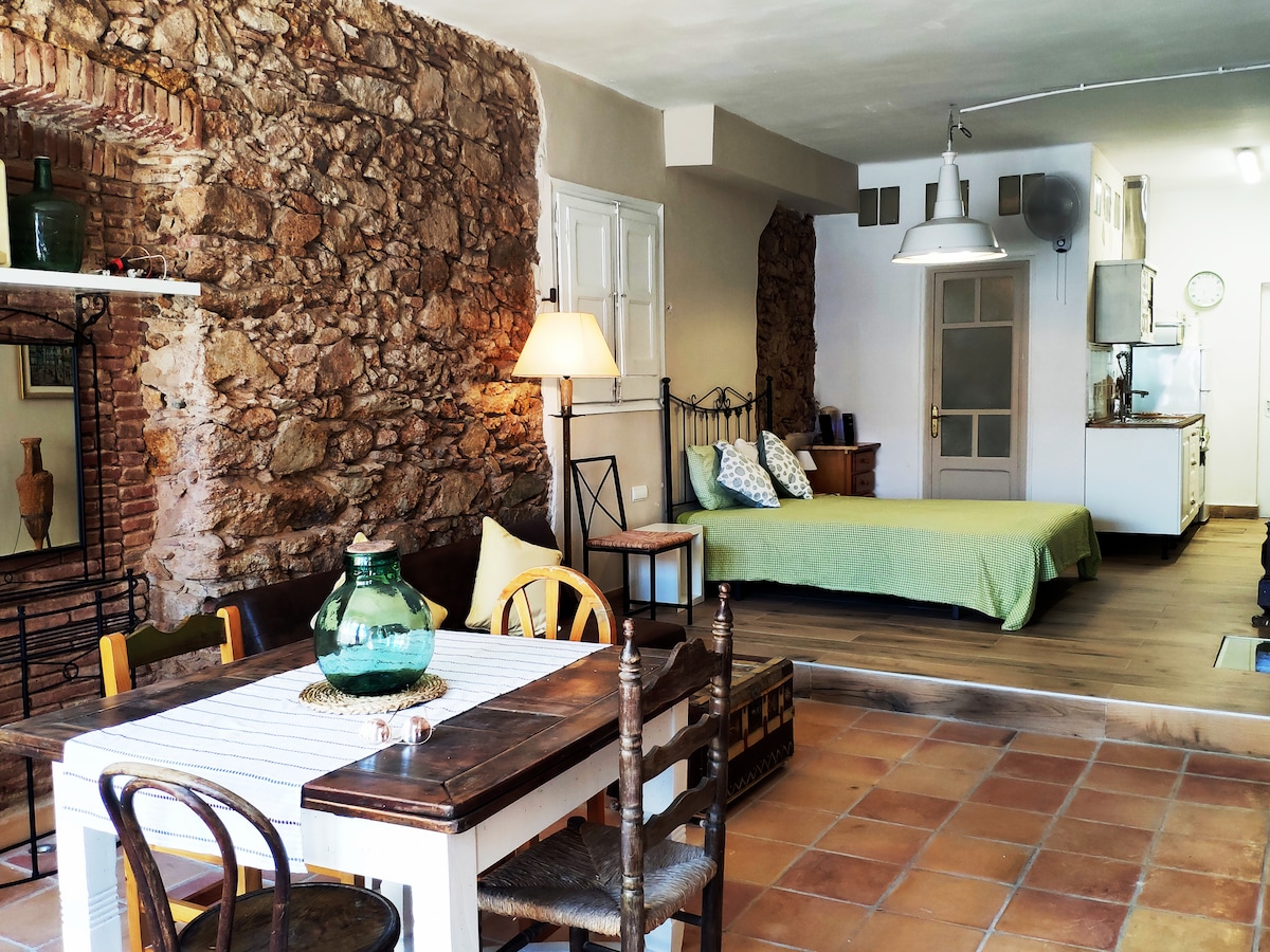 A cozy loft design is featured with exposed stone walls and wooden accents. The arrangement includes a dining table and chairs, along with a spacious bed adorned in light green bedding. A well-equipped kitchen is visible in the background, contributing to a functional living space.