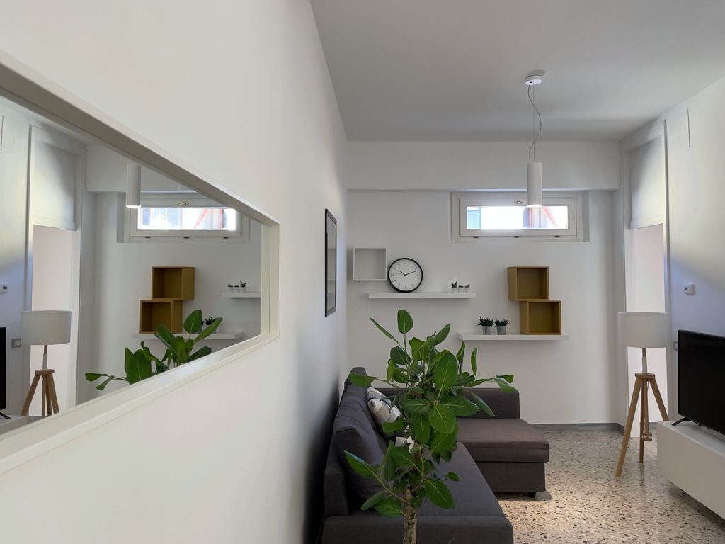 A modern living area features a dark gray sectional sofa positioned against a white wall. A plant adds a touch of greenery, while a minimalist clock and shelves with decorative items are mounted above a flat-screen television. Two windows allow natural light to brighten the space.