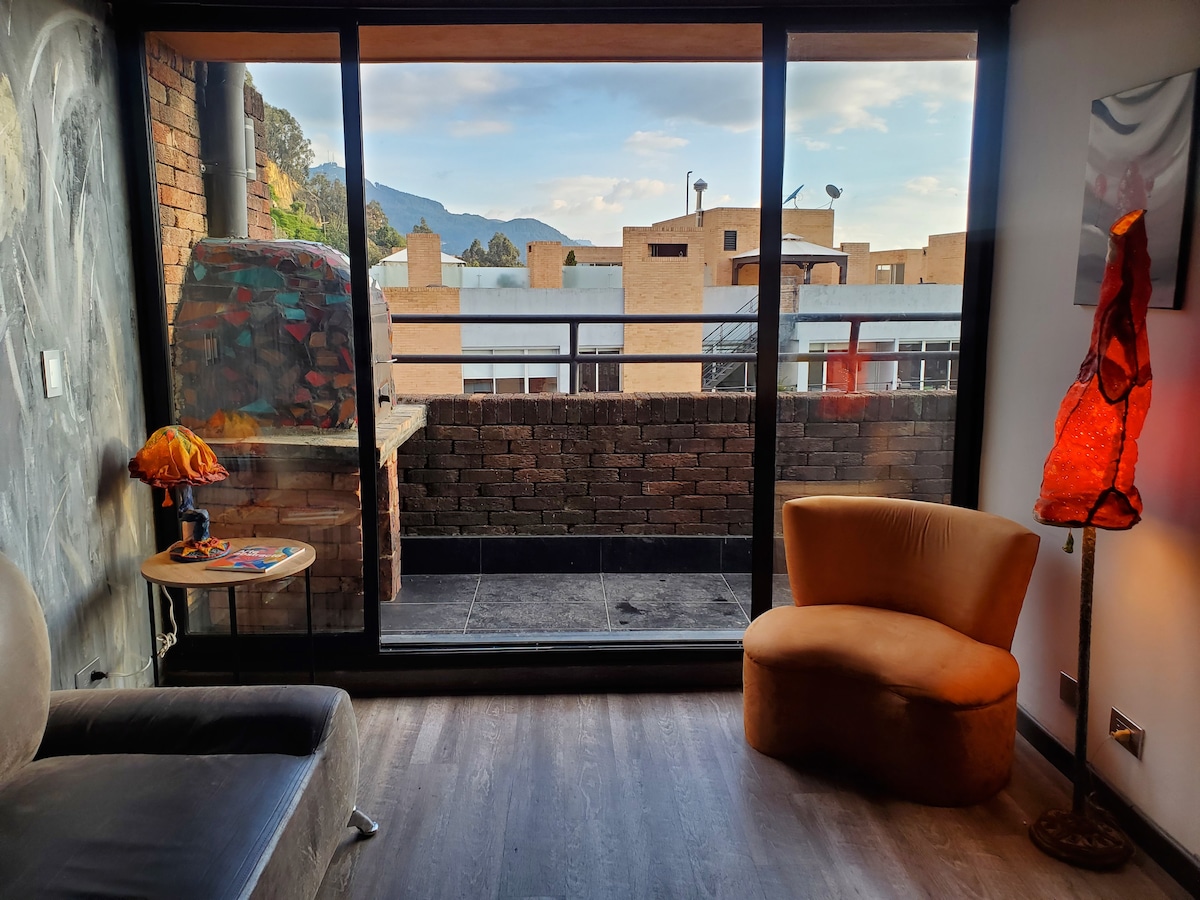 A seating area features a plush, curved chair beside a small table. Large glass doors offer a view of the outdoor terrace, with mountains visible in the distance. A colorful lamp and textured wall add visual interest to the space.