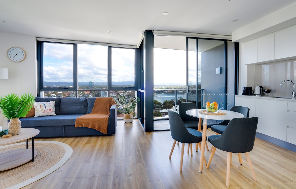 A bright living space features a comfortable couch with a cozy throw, complemented by a round dining table and chairs. Large windows allow for natural light and offer views of the surrounding landscape, highlighting the connection to the outdoors.