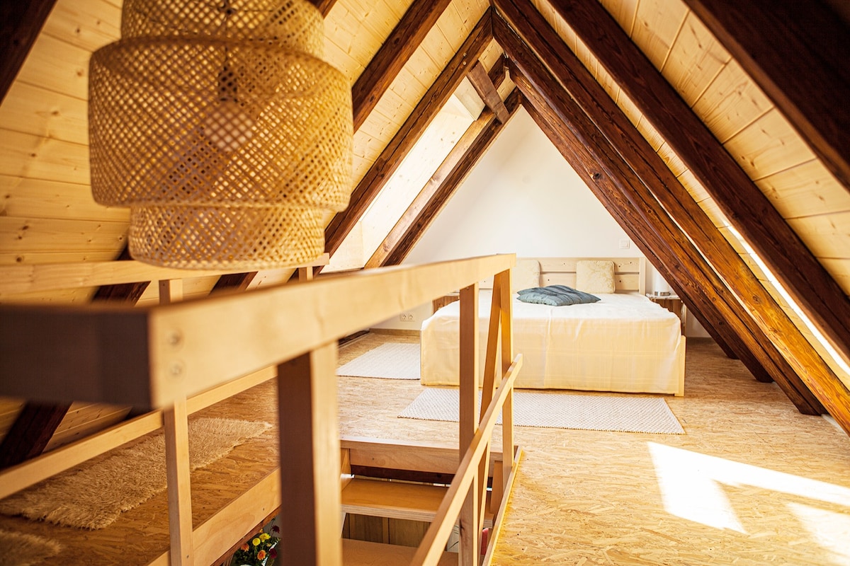 An airy attic room features sloped wooden ceilings with exposed beams. A spacious bed is positioned centrally, complemented by natural wood accents. A woven light fixture hangs overhead, and a soft area rug is placed on the floor, enhancing the cozy ambiance. Large windows allow ample natural light.