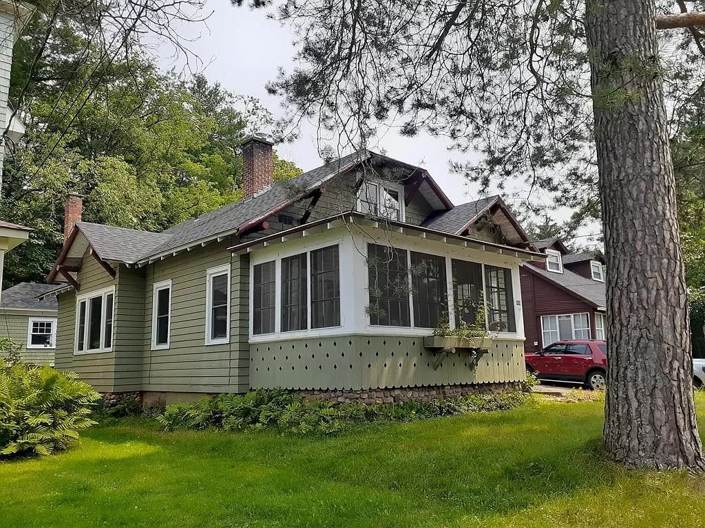 A charming house with a green exterior showcases a three-season porch featuring large windows, surrounded by landscaped grass and trees. The roof is pitched, with a brick chimney visible. Nearby, a red house and a parked vehicle add context to the peaceful neighborhood setting.