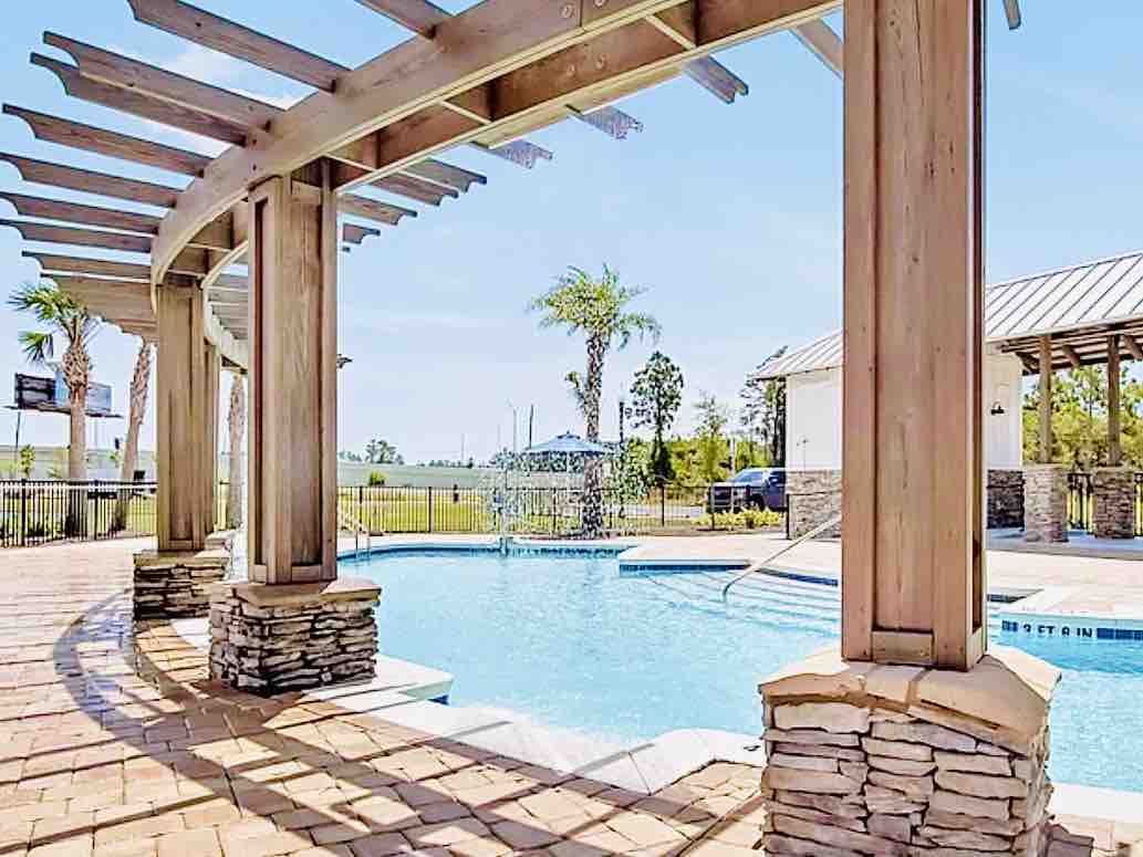 A community pool area is framed by wooden beams, highlighting a refreshing swimming pool with gentle waves. Surrounding greenery, including palm trees, provides a serene backdrop, while the stone pillars add a rustic touch to the welcoming space.