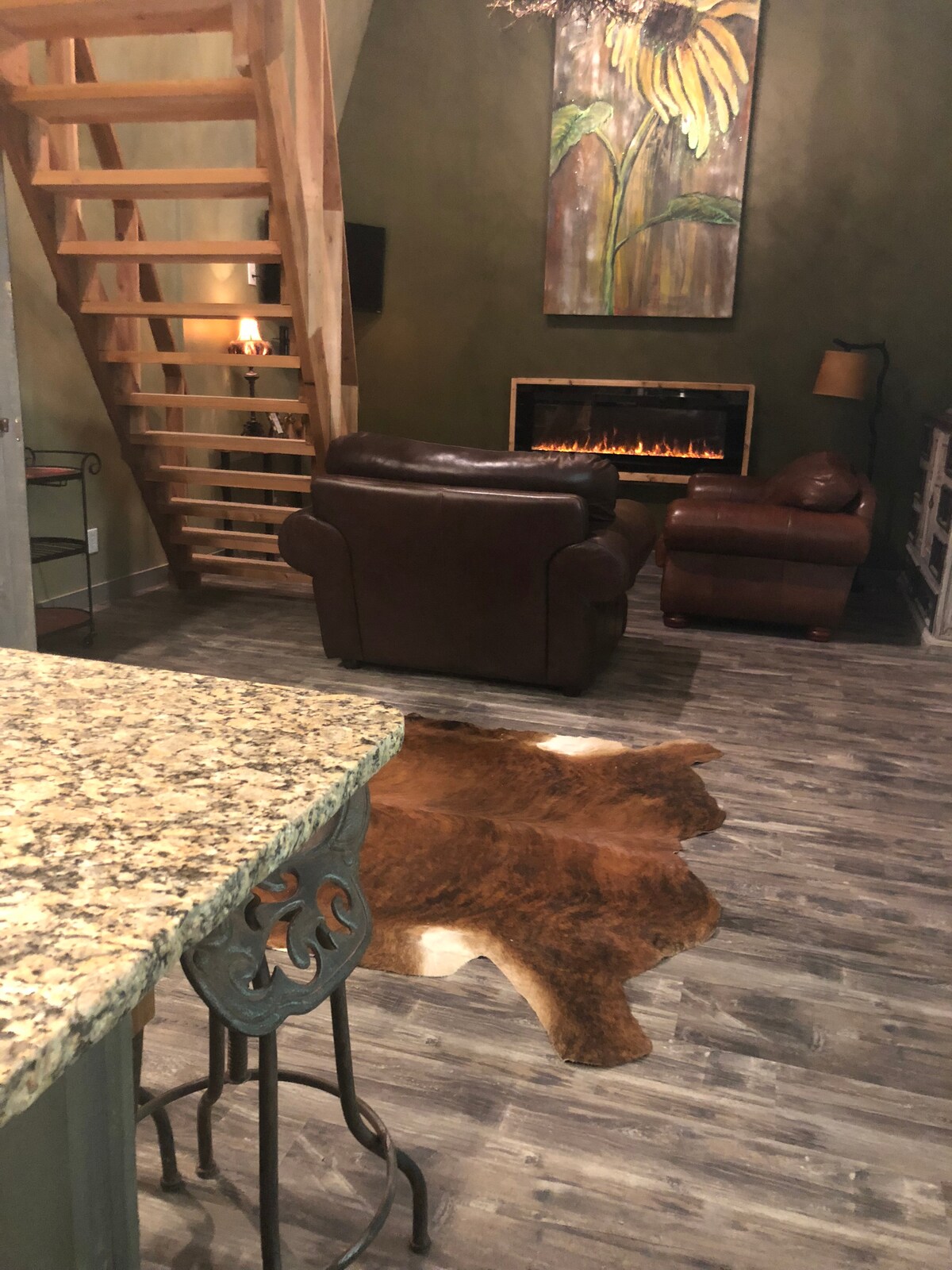 An open living area features a cozy leather loveseat and a large chair positioned near a modern electric fireplace. A rustic area rug lays on the wood floor, adding warmth to the space. A staircase leads to the upper bedroom, with a decorative wall panel above.