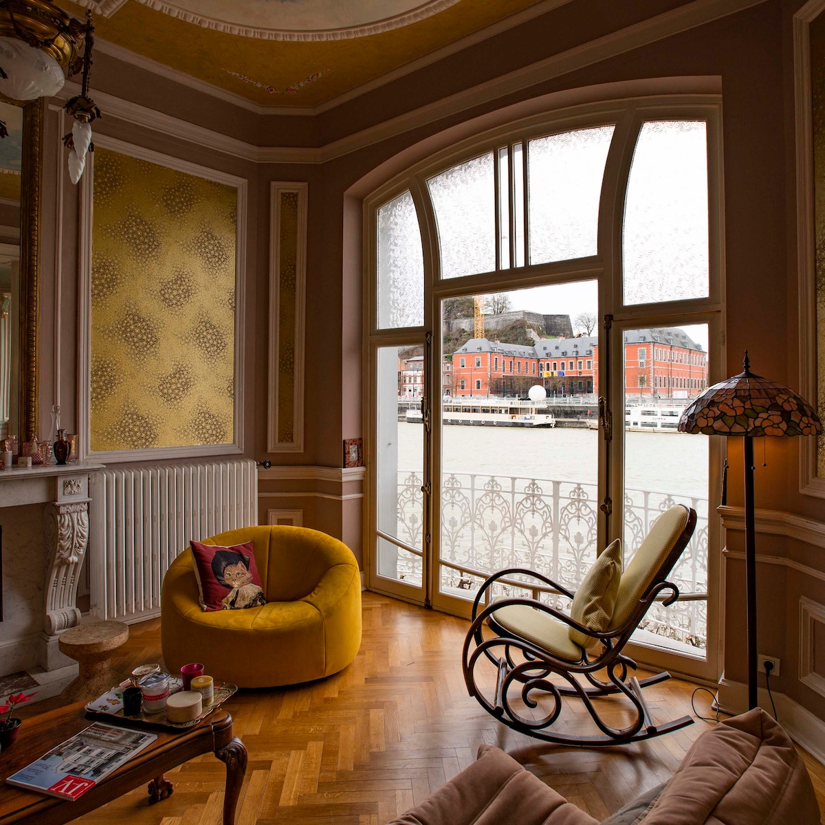 A cozy seating area features a yellow armchair and a rocking chair positioned by large windows, allowing natural light to fill the space. The view showcases the river and historic buildings across the water, enhancing the room's inviting ambiance.