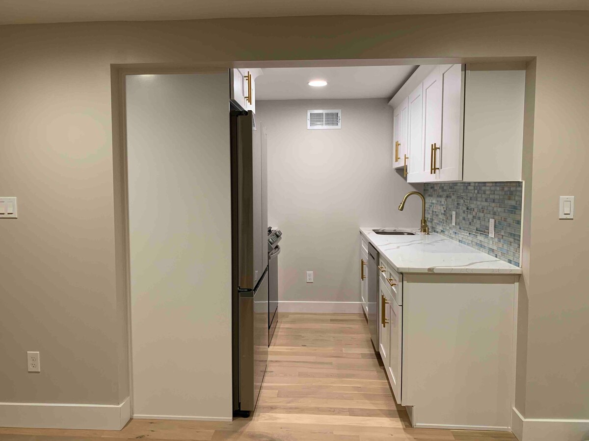The kitchen features modern white cabinetry and sleek gold accents, complemented by a mosaic tile backsplash. Stainless steel appliances, including a refrigerator and gas range, are present. Ample counter space is available for meal preparation, illuminated by overhead lighting.