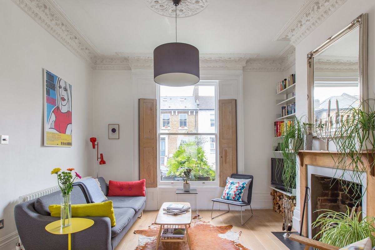 A modern living area features mid-century furnishings including a gray sofa with colorful cushions. A bright yellow side table holds a vase of flowers. Large sash windows invite natural light, and a decorative fireplace adds character, while built-in shelves display books and decorative items.