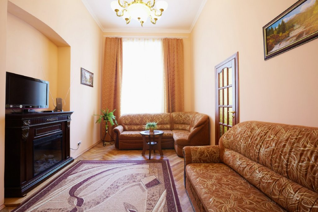 A spacious living area is featured, with two cozy sofas arranged around a small wooden table. Natural light filters through large windows, complemented by elegant drapes. A television is positioned on a dark wooden cabinet, and a decorative painting adds a touch of character to the wall.