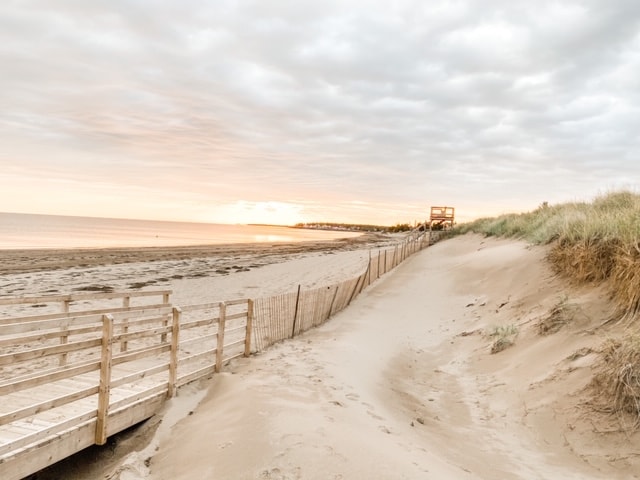 A sandy pathway leads to a tranquil beach at sunset, with gentle waves reflecting soft hues in the sky. A wooden boardwalk offers access to the water, while grass-covered dunes frame the scene, enhancing the serene coastal atmosphere.
