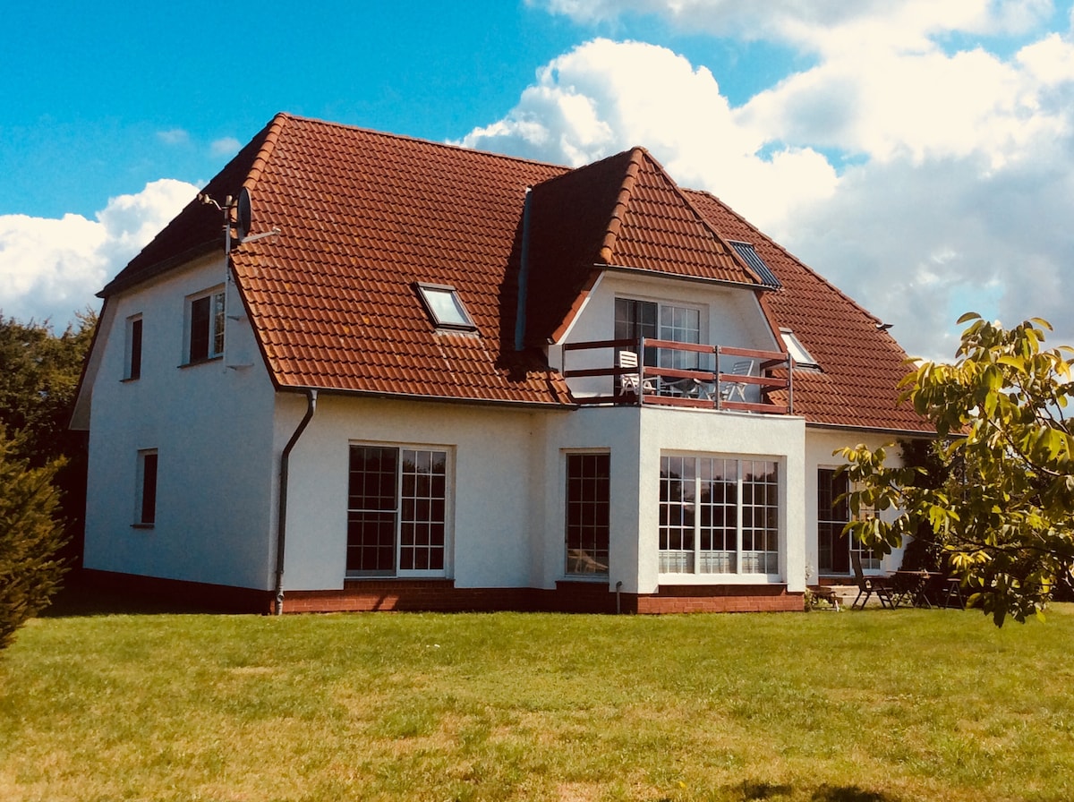 A spacious two-story house is showcased, featuring a red-tiled roof and multiple large windows. A balcony extends from the upper level, while a green lawn surrounds the exterior. Soft clouds drift across a clear blue sky, enhancing the inviting atmosphere of the property.