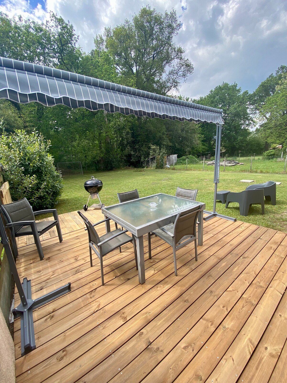 A wooden deck is furnished with a glass table surrounded by four chairs, offering a space for dining. A black grill is positioned nearby. An awning provides shade above, while green trees and an open lawn can be seen in the background.