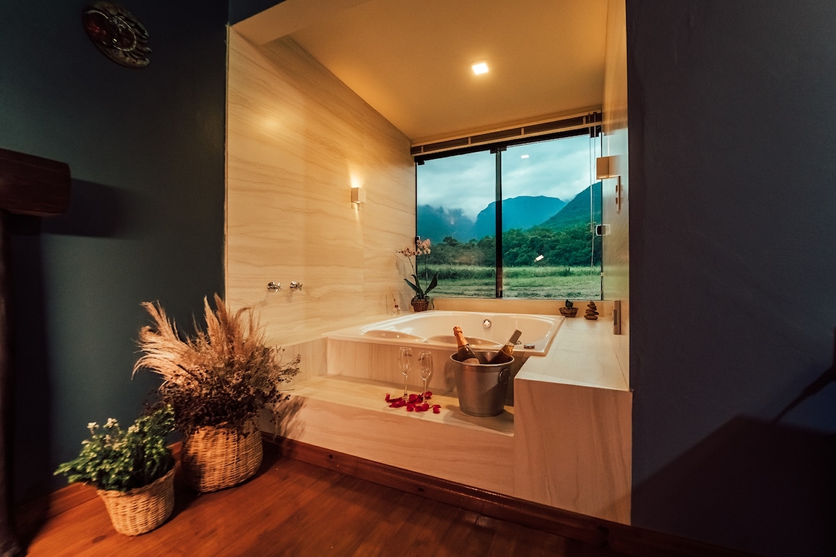 A modern bathroom features a spacious whirlpool tub positioned near a large window. Scenic views of rolling hills and nature are visible beyond the glass. Elegant lighting and tasteful decor complement the serene space, enhancing the atmosphere for relaxation.