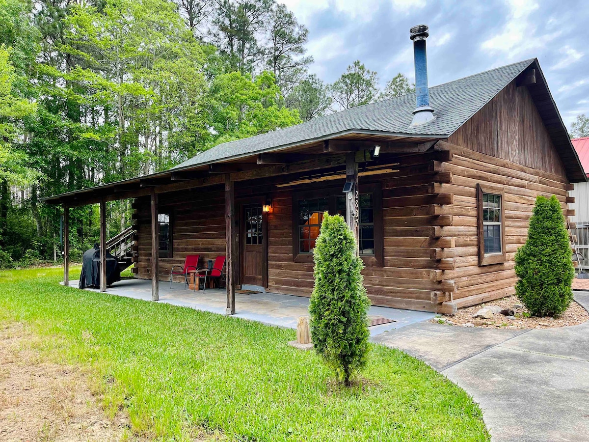 The exterior of a log cabin is shown, featuring a covered porch with seating. A chimney can be seen on the roof. Surrounding greenery adds a natural touch, while the cabin's wooden structure showcases its rustic charm.