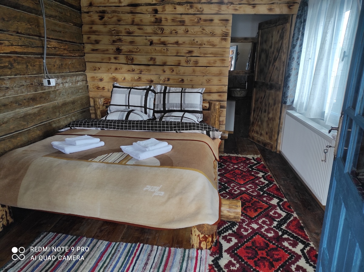 A rustic wooden bedroom features a spacious bed adorned with a checkered blanket and neatly arranged towels. The room is complemented by a traditional patterned rug and wooden accents, with natural light filtering through a large window covered by sheer curtains.