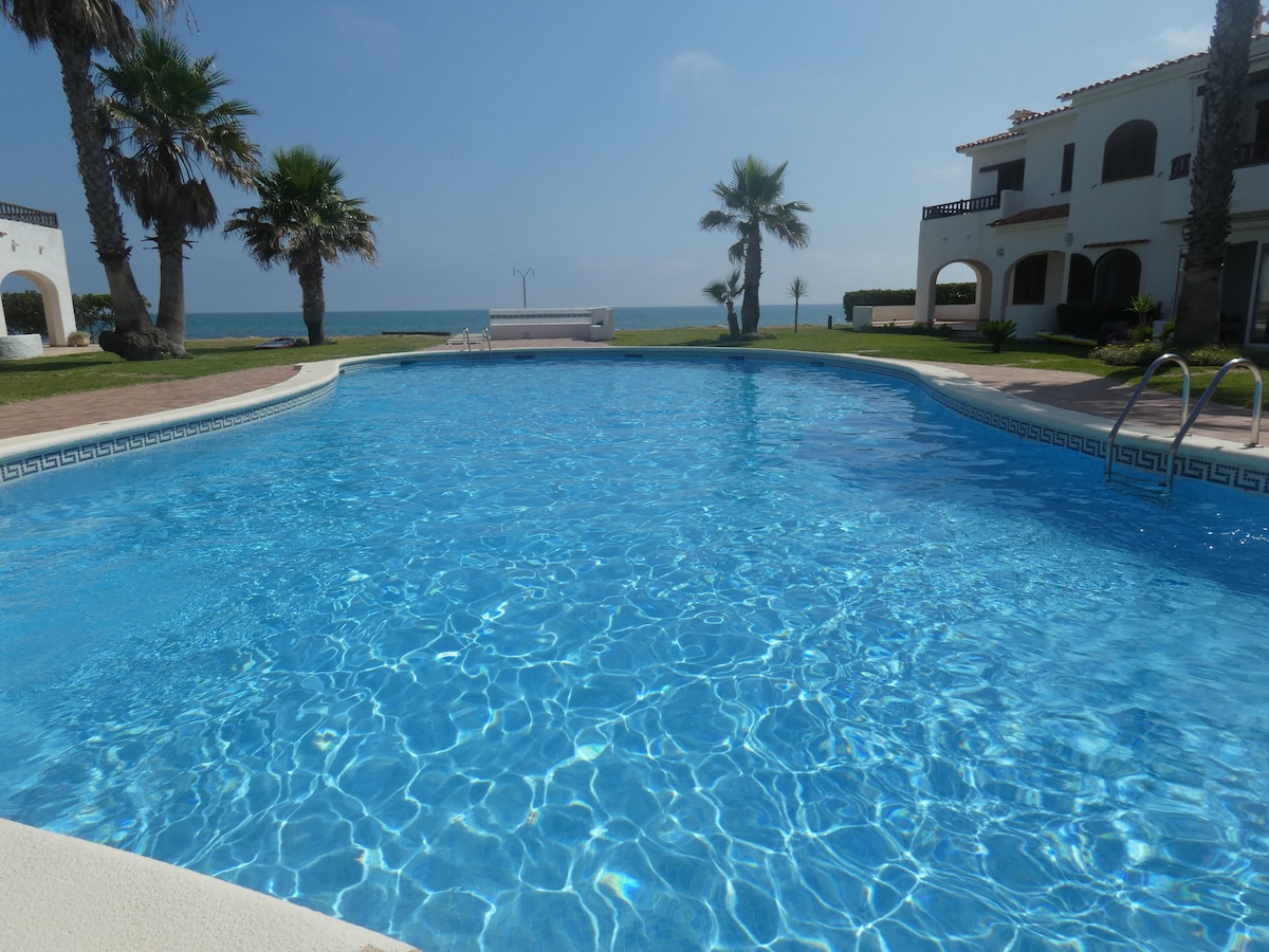 A clear blue swimming pool is framed by lush greenery and palm trees, offering a view of the sea in the background. Sunlight reflects on the water's surface, creating a sparkling appearance. The pool area is bordered by a well-maintained garden.