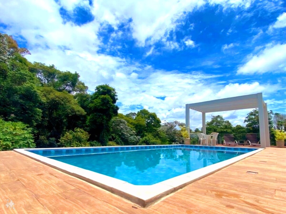A heated swimming pool is surrounded by a wooden deck and lush greenery. The clear blue water reflects the bright sky and scattered clouds. A shaded area with chairs is visible nearby, offering a comfortable space to relax and enjoy the natural surroundings.