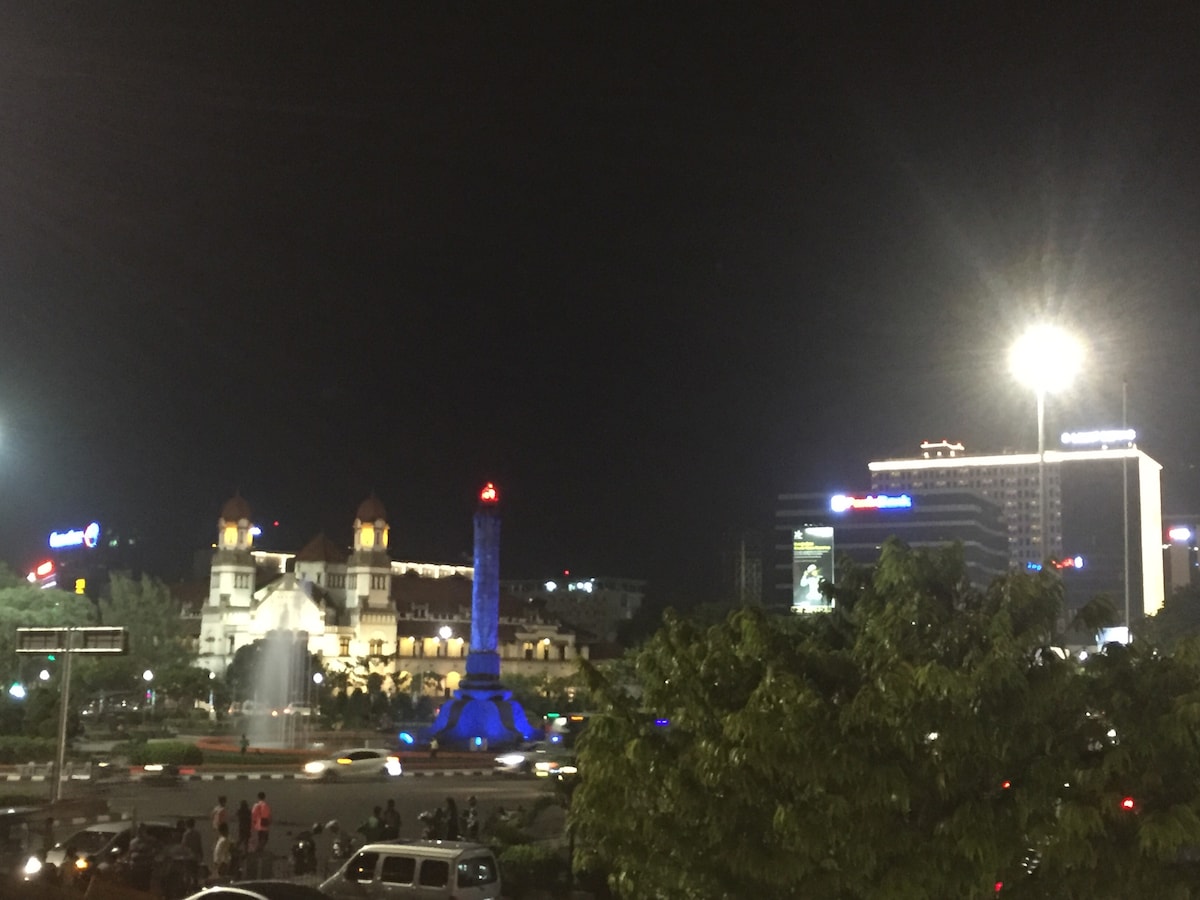 A night view of Tugu Muda, the iconic monument of Semarang, is illuminated with blue lights, surrounded by bustling traffic. Modern buildings and bright storefronts contribute to the vibrant atmosphere, while greenery is visible in the foreground, providing a contrast to the urban scene.