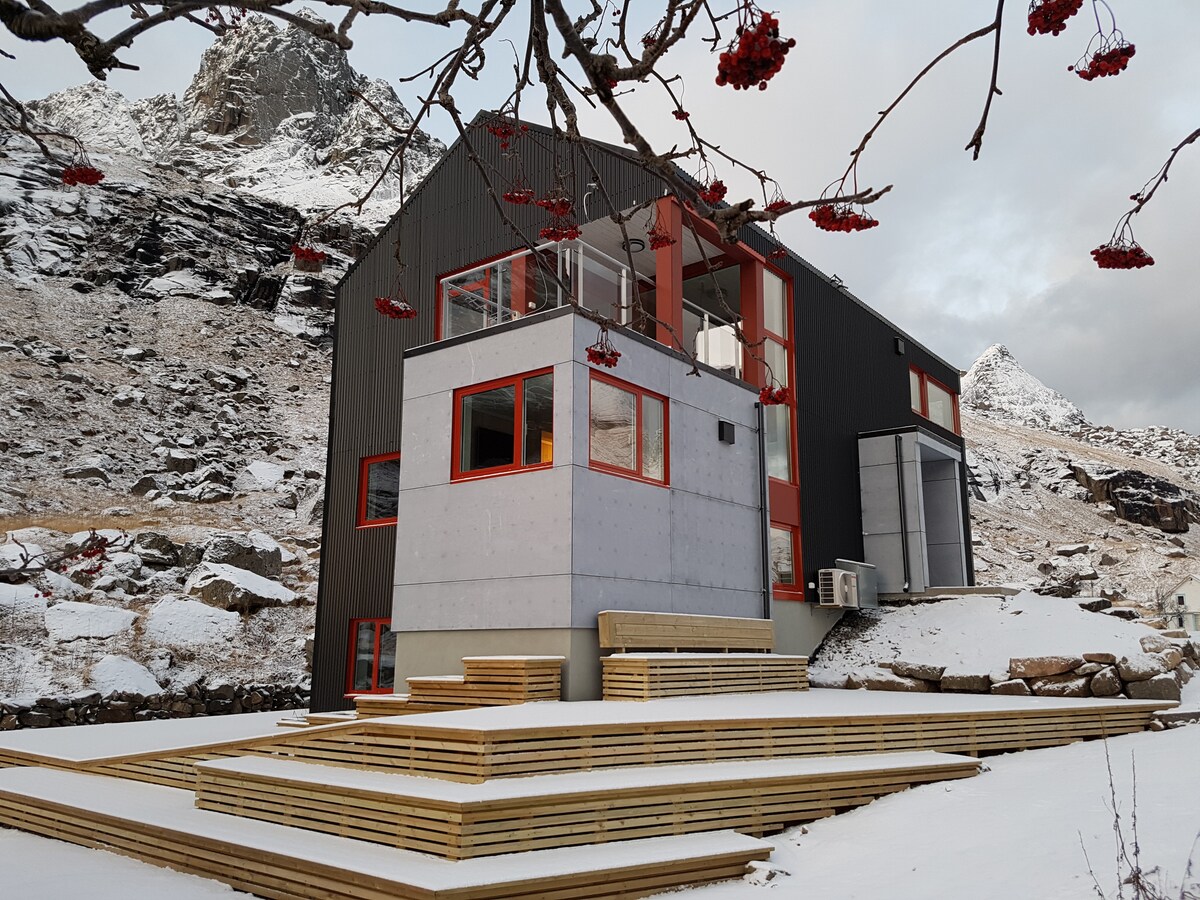 A modern house is set against a snowy landscape and rugged mountains, featuring a combination of concrete and dark wood exteriors. A spacious wooden deck with multiple levels provides outdoor seating options, while large windows offer views of the surrounding nature.