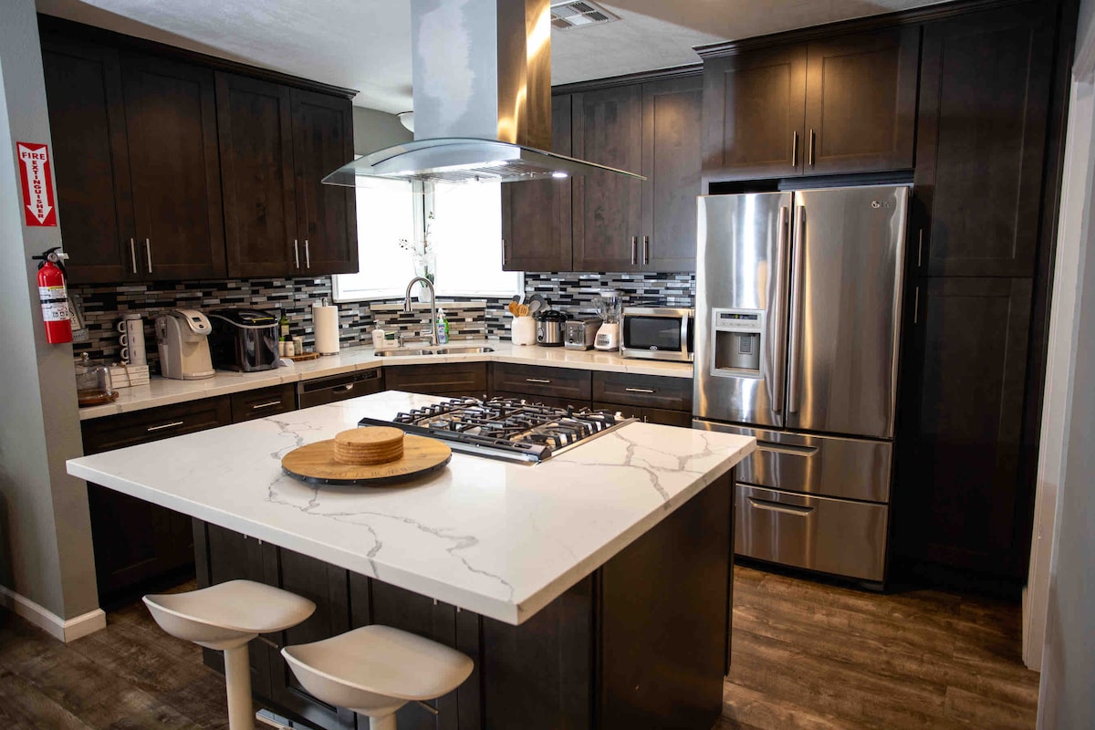 The modern kitchen features dark cabinetry with stainless steel appliances, including a large refrigerator and built-in oven. A central island with a marble countertop and three barstools offers ample space for dining or food preparation. A stylish backsplash complements the overall design.