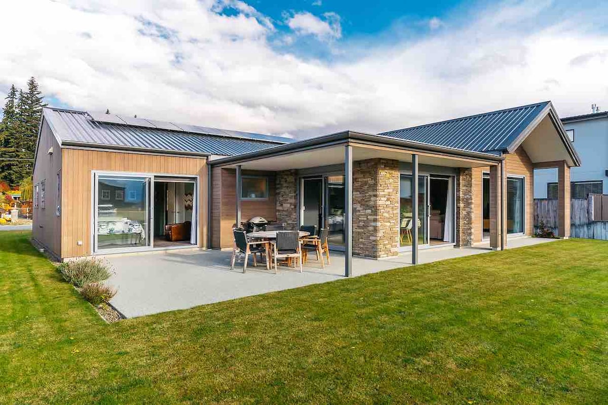 The exterior of a modern house is showcased, featuring a covered patio area with outdoor furniture. Large windows provide views into the spacious interior. A lawn with soft landscaping complements the structure, while the slate roof and stone accents enhance the contemporary design. Mountain views are visible in the background.