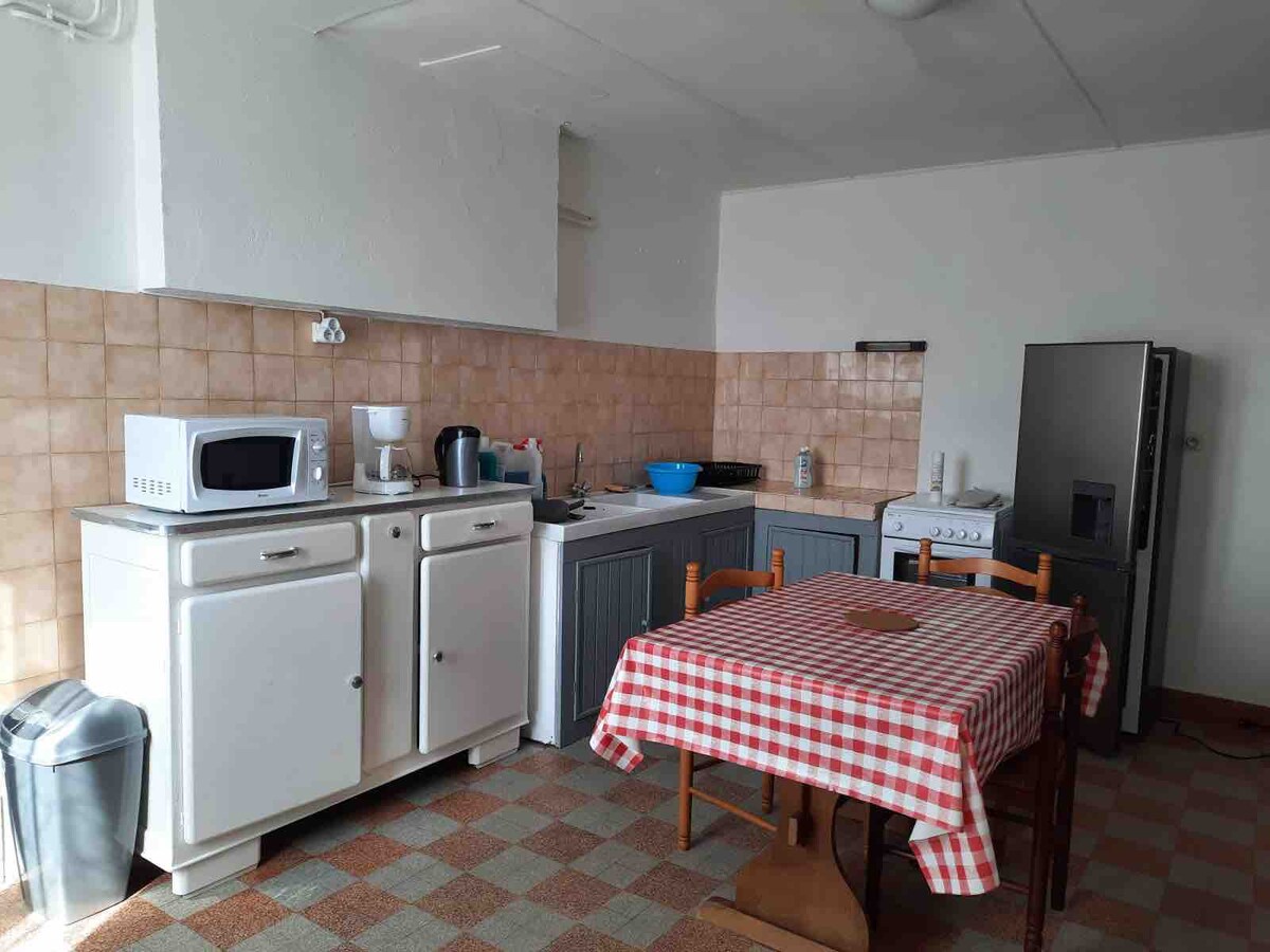 A functional kitchen area features a tiled backsplash with beige and brown tones. A red and white checkered tablecloth covers a wooden table surrounded by four chairs. Appliances include a microwave, coffee maker, and a large refrigerator, with ample counter space for meal preparation.