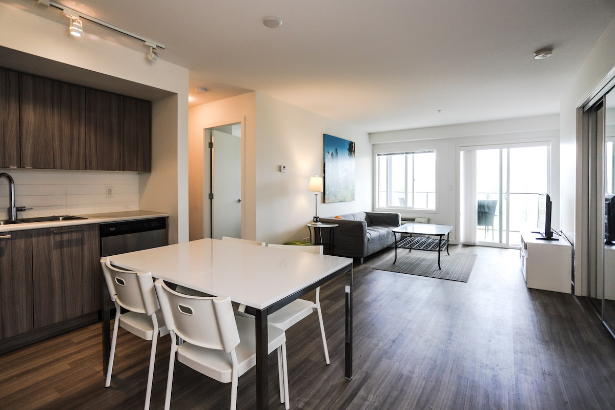 A spacious living area features large windows that provide natural light and views of the balcony. The modern kitchen is equipped with sleek cabinetry and necessary appliances. A dining table with four chairs is positioned adjacent to a comfortable seating arrangement, with a television mounted nearby.