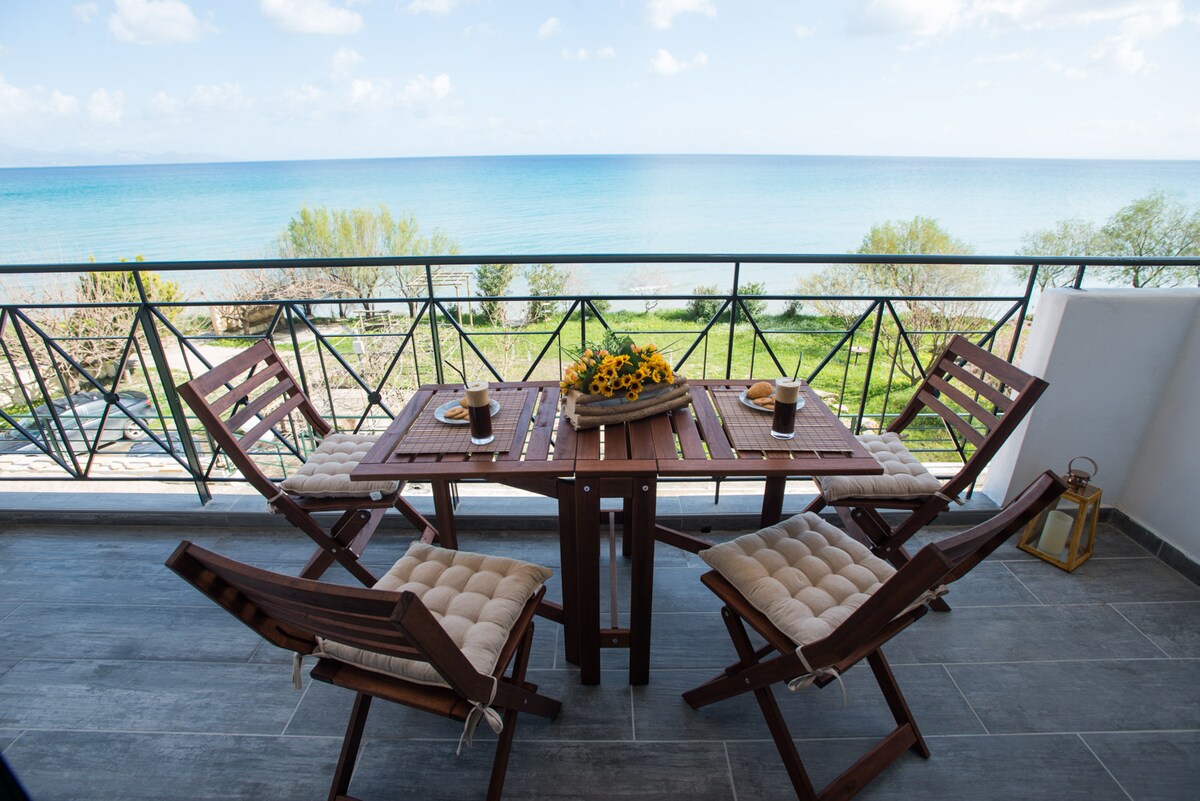 A spacious outdoor terrace is furnished with a wooden dining table and four folding chairs, showcasing a view of the tranquil sea. Sunflowers are arranged on the table, complemented by two drinks, with the blue waters extending towards the horizon.