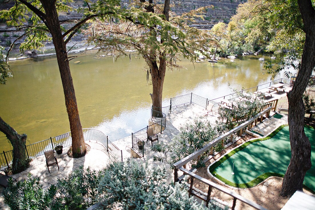A view of the Guadalupe River features a custom stone patio with a concrete bulkhead. Lounge chairs are positioned along the riverfront, and a two-tier common area includes a putt-putt green. Lush greenery surrounds the space, enhancing the natural setting.