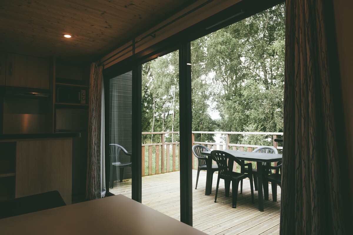 A view from inside the cottage shows large sliding glass doors opening to a spacious terrace. The terrace features a set of outdoor chairs and a table, surrounded by greenery. Natural light floods the interior, highlighting wooden accents.