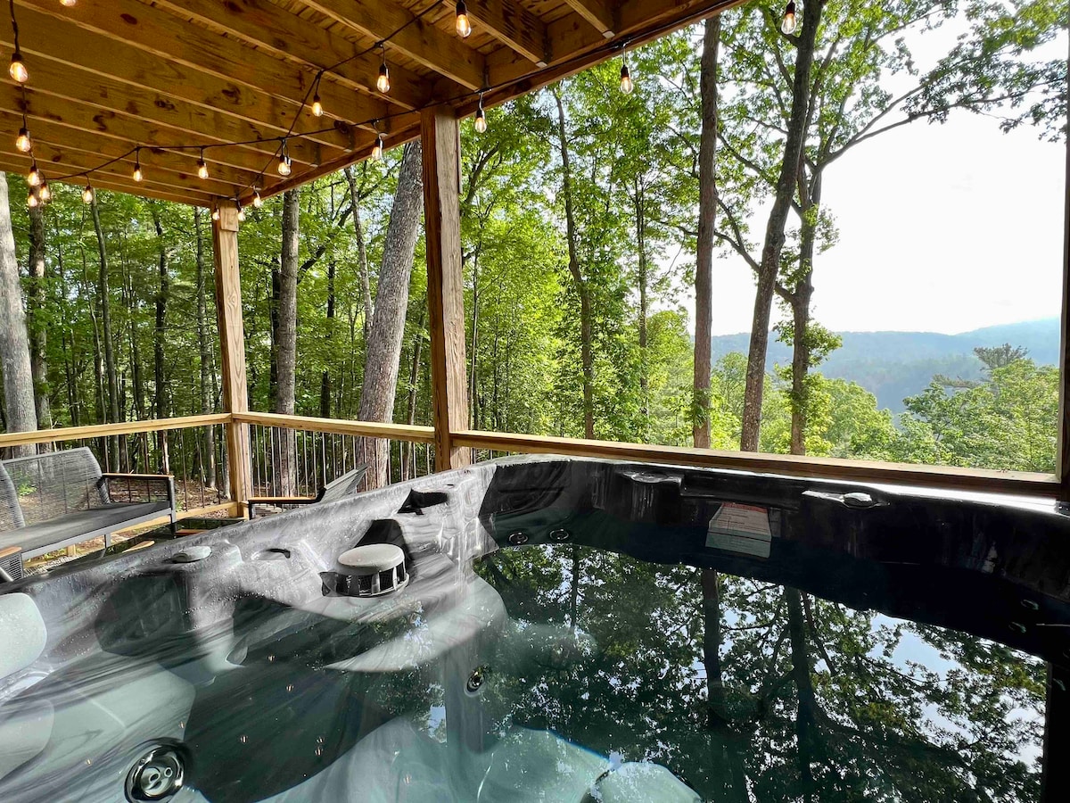 A jetted hot tub is situated on a covered deck, surrounded by a wooden railing. Trees create a natural backdrop, with expansive mountain views visible beyond. Warm lighting from string lights adds ambiance to the outdoor space.
