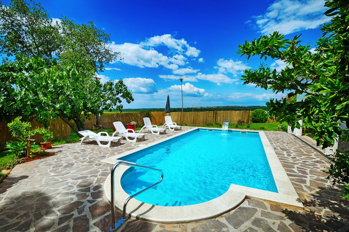 A spacious outdoor pool is surrounded by a stone patio, furnished with several white lounge chairs under a clear blue sky. Lush greenery and trees offer natural shade, while a lamp post stands by the pool area, enhancing the serene atmosphere of the surrounding landscape.