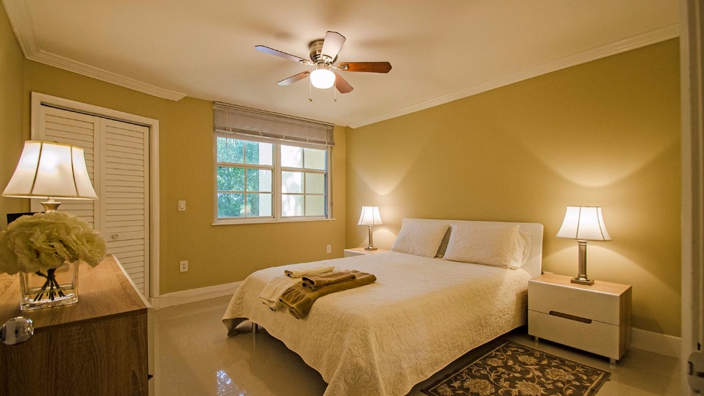 A well-lit bedroom features a spacious bed with light bedding, flanked by two bedside lamps on wooden nightstands. A ceiling fan is positioned above, and a window allows natural light to fill the room. A decorative rug adds a touch of warmth to the polished floor.