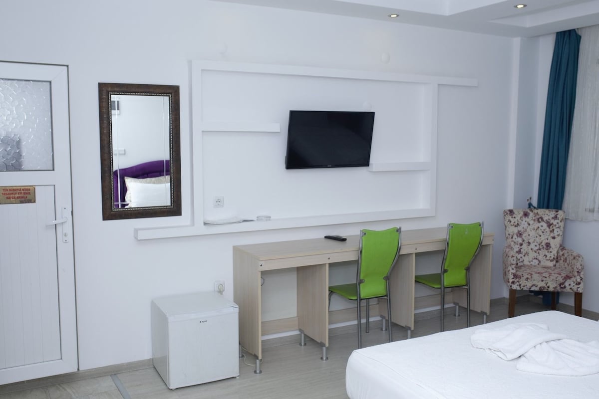 A modern room features a wall-mounted television above a simple desk with two green chairs. A compact refrigerator is positioned nearby. Soft light is provided by overhead fixtures and windows adorned with blue curtains, enhancing the bright and airy feel of the space.