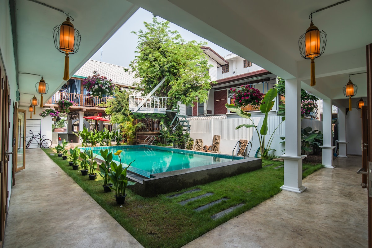 A serene courtyard features a central swimming pool surrounded by lush greenery and ornamental plants. Lanterns hang from the ceiling, casting a warm glow. Nearby, traditional Thai-style buildings and a bike are visible, contributing to the tranquil ambiance of the space.