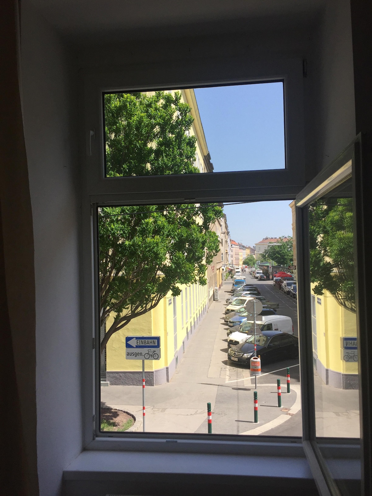 A view from a window showcases a tree-lined street with colorful buildings in the background. Numerous parked vehicles line the street, and a clear blue sky is visible, contributing to a bright and open atmosphere.