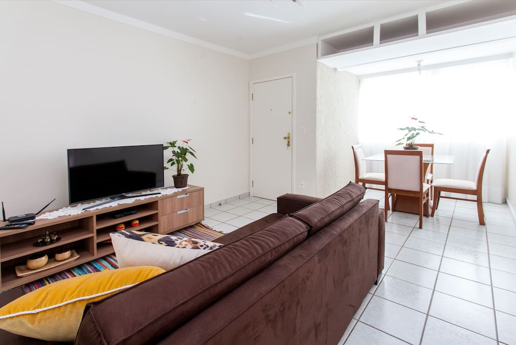 Vista da sala de estar e da sala de jantar, com mesa e quatro cadeiras. Ao fundo, a porta de entrada do apartamento.