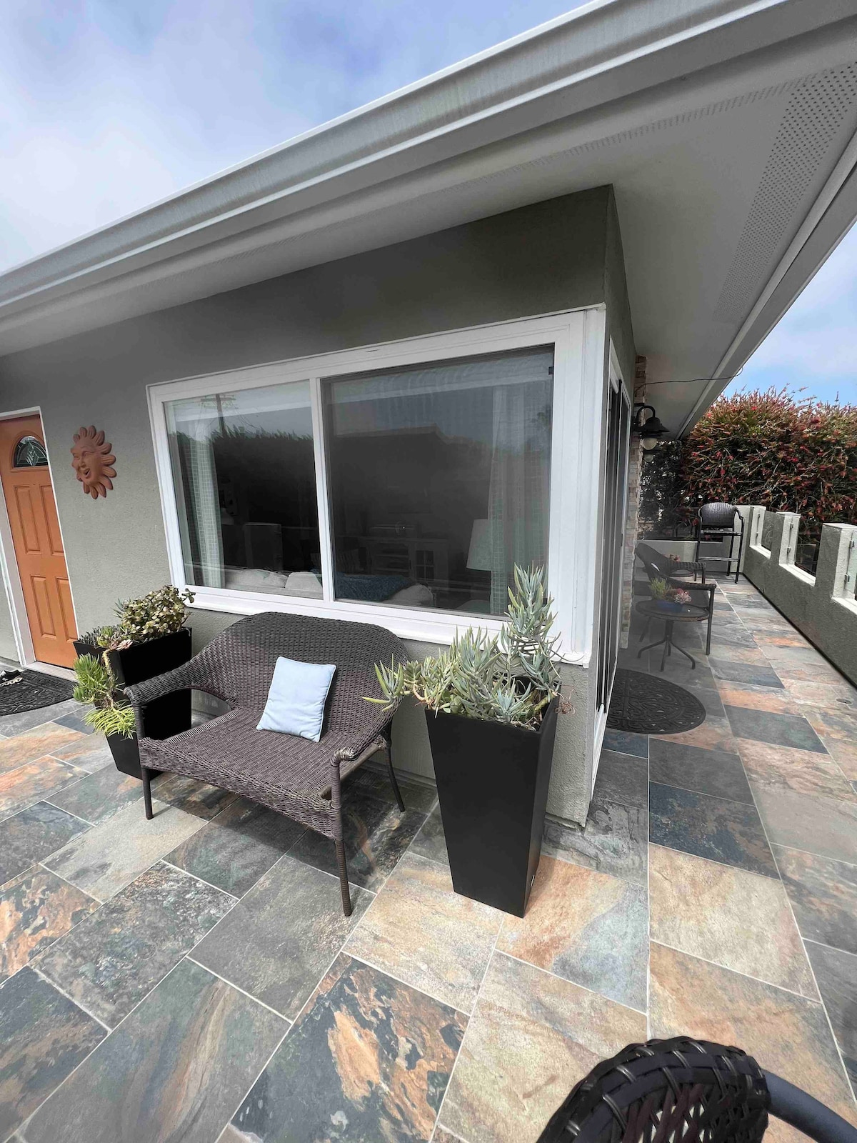 A welcoming patio area is featured with a woven seating arrangement, complemented by potted plants. Natural stone tiles create a textured surface, while a large window provides a view of the surroundings, enhancing the connection to the outdoor space.